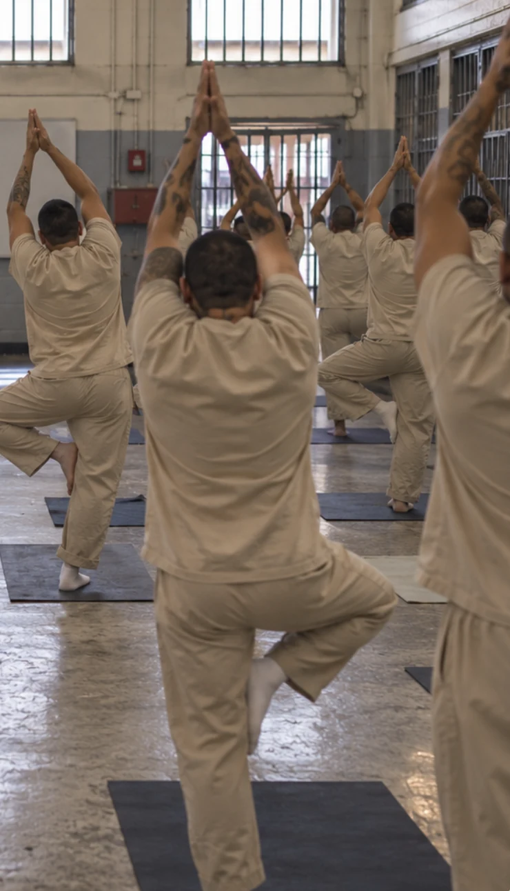 Clase de yoga en un salón de prisión: personas de espaldas en postura del árbol, con las manos juntas sobre la cabeza.