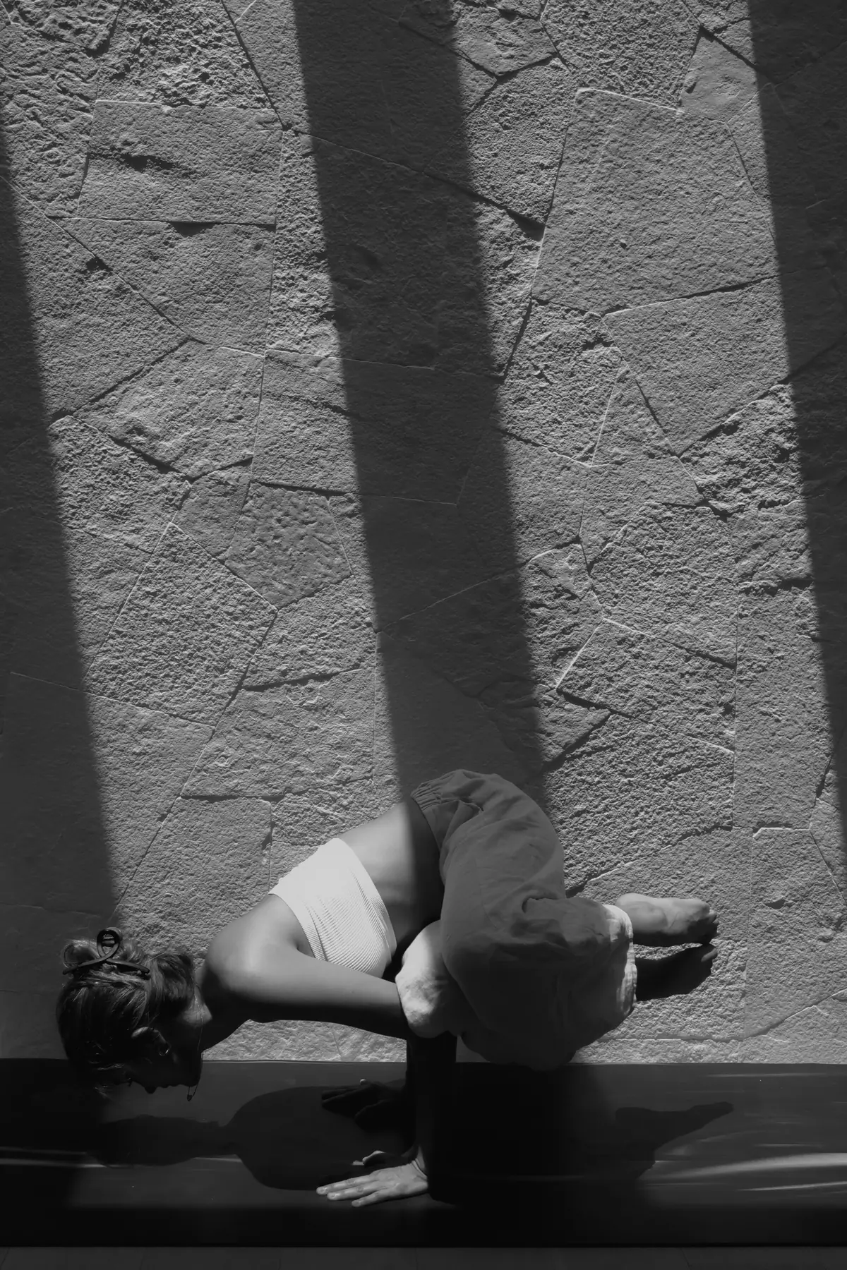 Mujer en postura de cuervo frente a un muro donde la luz dibuja sombras de barrotes.